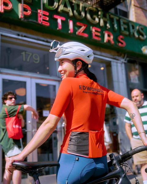 Woman in EdW Edition Jersey, Cinnabar Orange, smiling on her bike outside a restaurant, showcasing stylish and functional cycling apparel.